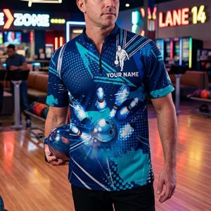 Könnte beinhalten: Ein blaues Bowling-Shirt mit einem Design aus Kegeln und Kugel. Das Shirt hat einen Reißverschluss und den Text "YOUR NAME" aufgedruckt. Das kurzärmelige Shirt hat ein Bowling-Thema.