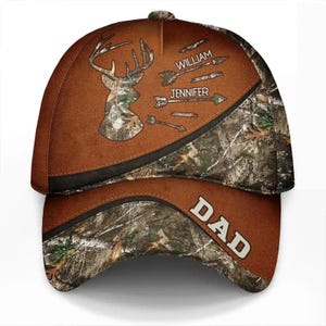May include: A baseball cap featuring a brown and camouflage pattern. The front displays a deer silhouette, two arrows with the names WILLIAM and JENNIFER, and the word DAD printed in white on the brim. The design includes dark brown accents.