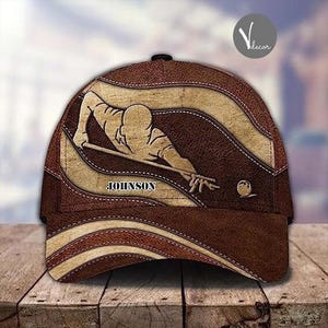 Könnte beinhalten: Braune und beigefarbene Baseballkappe mit einem braunen Lederdesign. Die Kappe zeigt eine Grafik einer Person, die Billard mit einem Queue und einer Billardkugel spielt. Der Text "JOHNSON" ist auf die Kappe gedruckt.