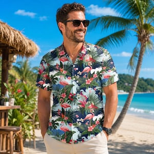 May include: A short-sleeved button-down shirt featuring a dark blue background with a repeating pattern of pink flamingos, green palm leaves, and red hibiscus flowers. The shirt has a classic collar and a straight hem, worn with light beige pants.