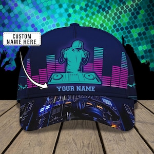 May include: A navy blue baseball cap with a teal and pink DJ graphic. The cap features the text "YOUR NAME" and "CUSTOM NAME HERE". The design includes sound wave bars and a DJ silhouette.