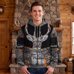 May include: A zip-up hoodie featuring a large deer head graphic on the chest, a forest camouflage pattern, and the text "Deer Hunter" printed on the front. The garment includes a hood with white drawstrings and a full-length front zipper closure.