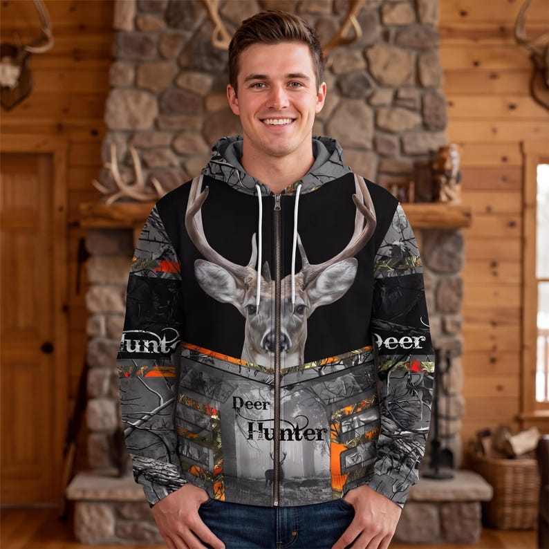 May include: A zip-up hoodie featuring a large deer head graphic on the chest, a forest camouflage pattern, and the text "Deer Hunter" printed on the front. The garment includes a hood with white drawstrings and a full-length front zipper closure.