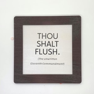May include: A square wall sign with a dark brown wood-grain frame and a white center panel. The text in black reads: THOU SHALT FLUSH. (The unwritten Eleventh Commandment). The sign features a clean, simple design with bold lettering.