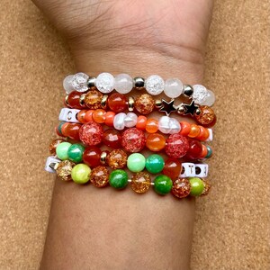 May include: A collection of beaded bracelets in various colors, including white, orange, green, and gold. Some bracelets feature star charms and letter beads. The bracelets are stacked on a wrist.