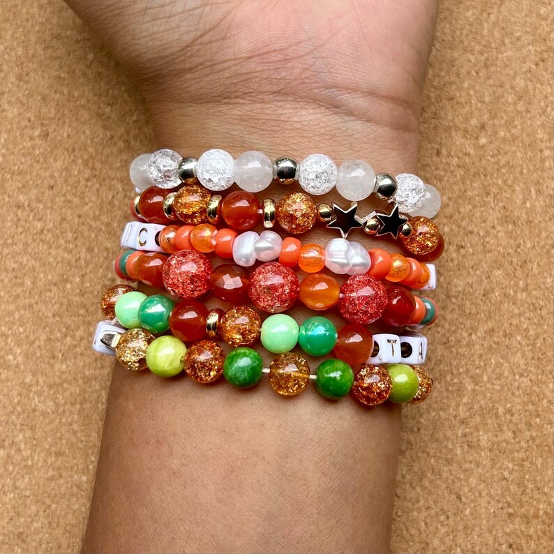 May include: A collection of beaded bracelets in various colors, including white, orange, green, and gold. Some bracelets feature star charms and letter beads. The bracelets are stacked on a wrist.