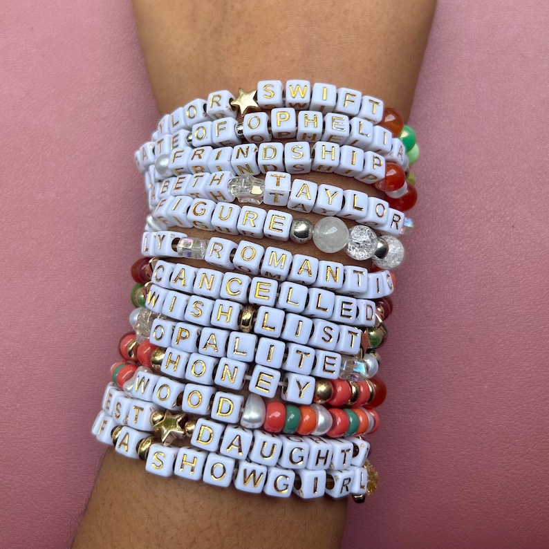 May include: A collection of beaded bracelets spelling out words like "SWIFT", "FRIENDSHIP", and "DAUGHTER" in gold lettering on white cubes. The bracelets are stacked on a wrist, with various colors and bead styles.