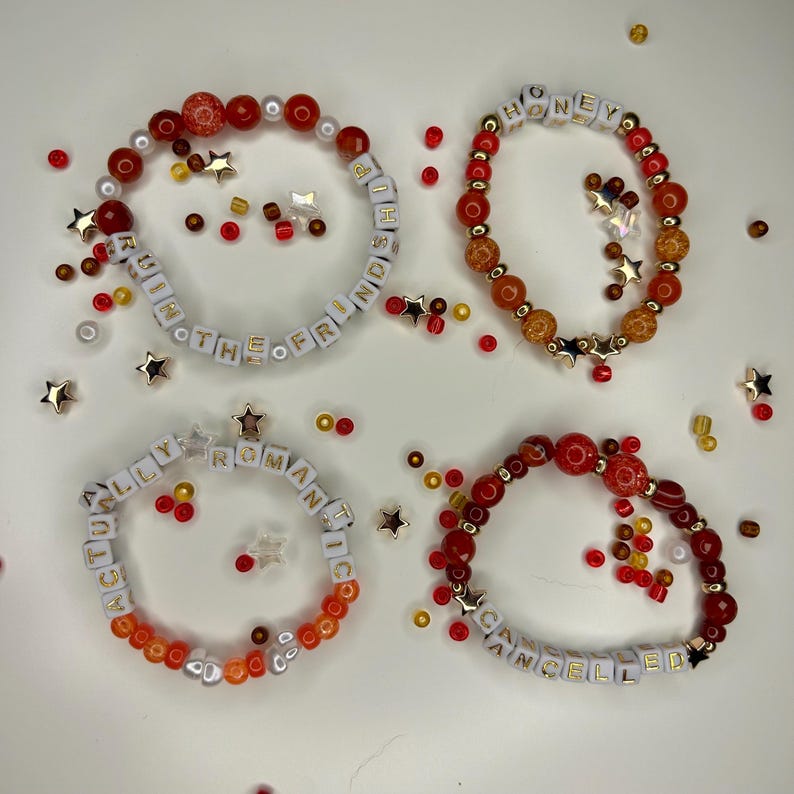 May include: Four beaded bracelets with white letter beads spelling out phrases like "ACTUALLY ROMANTIC" and "RUINING THE FRIENDSHIP". The bracelets are red, orange, and gold, with star and bead accents.