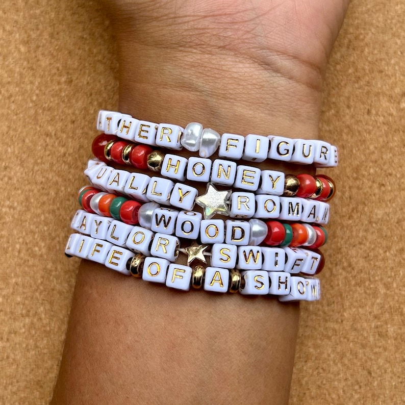 May include: A stack of white beaded bracelets spelling out phrases like "HER FIGURE", "HONEY", and "TAYLOR SWIFT". The bracelets feature gold and silver accents, with red, green, and orange beads interspersed throughout.
