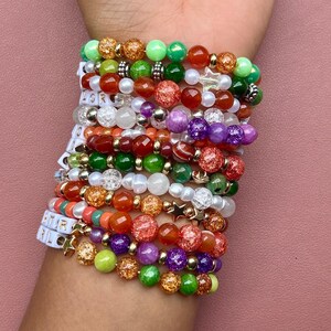 May include: A stack of beaded bracelets in various colors, including green, orange, red, purple, and gold. Some bracelets feature star-shaped charms and letter beads spelling out "STAR GIRL". The bracelets are worn on a wrist against a pink background.