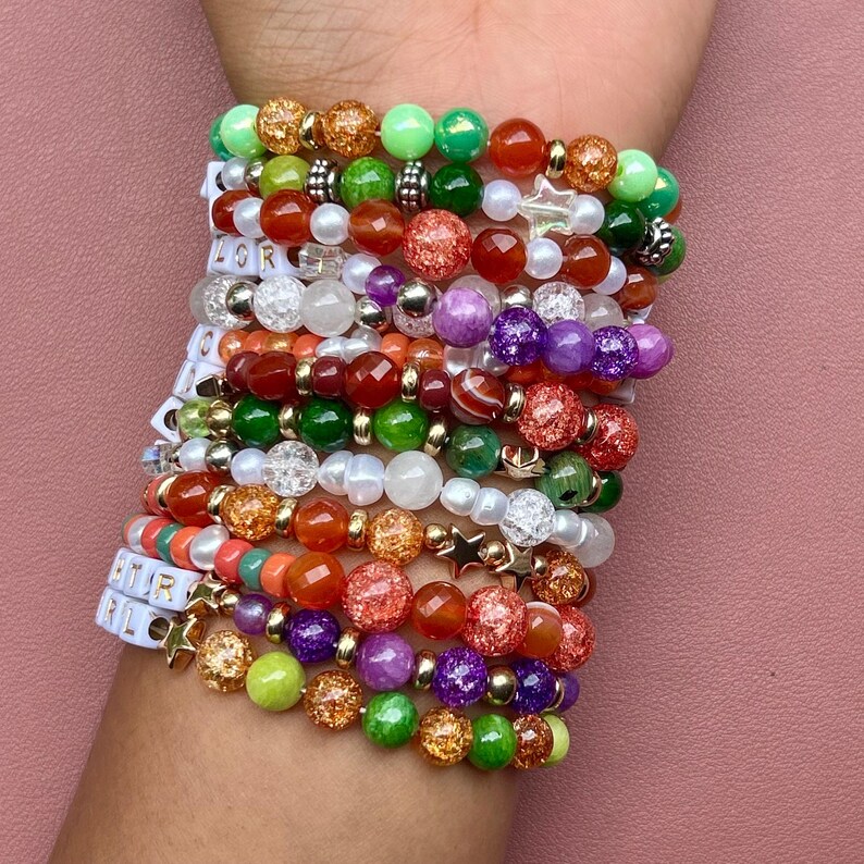 May include: A stack of beaded bracelets in various colors, including green, orange, red, purple, and gold. Some bracelets feature star-shaped charms and letter beads spelling out "STAR GIRL". The bracelets are worn on a wrist against a pink background.