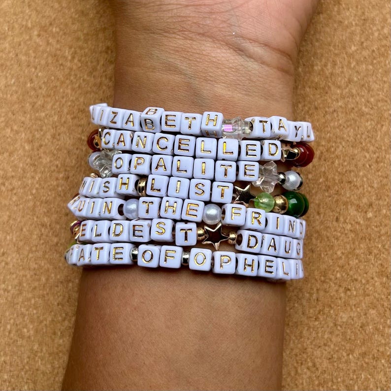 May include: A stack of white beaded bracelets with gold lettered words. The bracelets include the words: Elizabeth, Cancelled, Opalite, Wishlist, Win the, Eldest, and Fate of Ophelia. The bracelets are on a wrist.