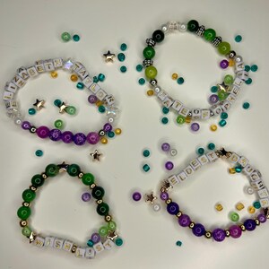May include: Four beaded bracelets with white letter beads spelling out names and words. The bracelets feature a variety of colorful beads in green, purple, and gold, with star-shaped accents. The bracelets are on a white surface.
