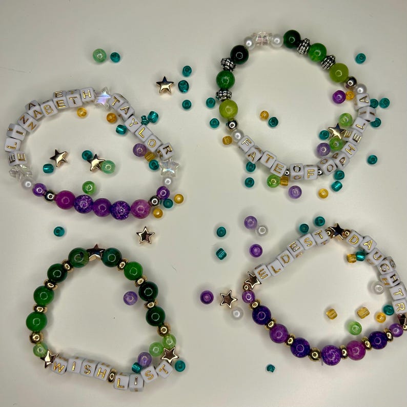 May include: Four beaded bracelets with white letter beads spelling out names and words. The bracelets feature a variety of colorful beads in green, purple, and gold, with star-shaped accents. The bracelets are on a white surface.