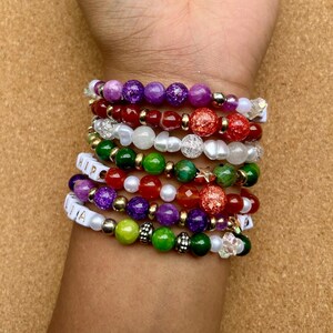 May include: A stack of beaded bracelets in various colors, including purple, red, green, and white. Some bracelets feature the text "HIP" and "LIA" on white letter beads. The bracelets are adorned with gold-colored spacers and decorative beads.