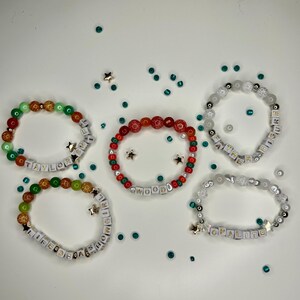 May include: Five beaded bracelets in various colors, including green, red, and white, with gold star charms and letter beads spelling out names and phrases. The bracelets are scattered on a white surface with scattered teal and gold star-shaped confetti.
