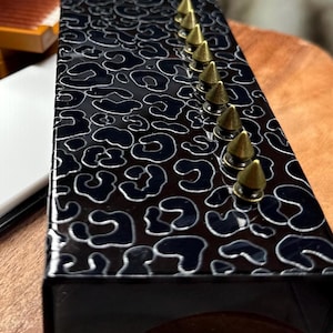 May include: A black rectangular box with a glossy leopard print design and a row of gold-colored cone studs. The box is set on a wooden surface, with a blurred orange box in the background.