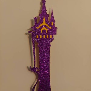 May include: Purple glitter paper cutout of a tower with a yellow silhouette of a castle on top. A silhouette of a person is climbing the tower.