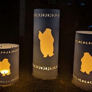 May include: Three cylindrical paper lanterns with a bear silhouette cutout and a crescent moon and star pattern. The lanterns are lit from within, casting a warm glow. The lanterns are dark blue.