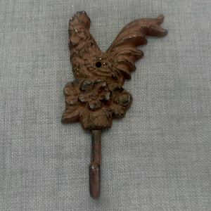 May include: A brown, cast metal wall hook featuring a detailed cockerel head and tail design positioned above a floral motif. The hook extends downwards from the base of the flower, and a small circular mounting hole is visible in the cockerel's neck.