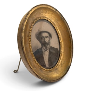 May include: An oval-shaped gold-toned picture frame with a decorative inner border, featuring a black and white portrait of a bearded person wearing a hat and a collared shirt. The frame is supported by a thin metal stand attached to the back.