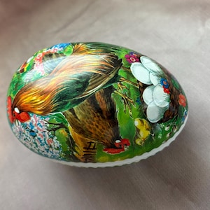 May include: An oval-shaped decorative egg with a detailed painting of a rooster and floral elements. The egg features a vibrant color palette of green, red, yellow, and blue. The rooster's feathers are depicted with intricate detail.