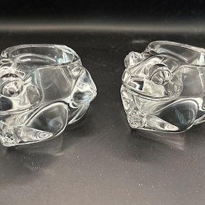 May include: Two clear glass frog-shaped candle holders. Each holder features a detailed frog face with bulging eyes and textured skin. The candle holders are designed to hold a small candle, adding a whimsical touch to any decor.
