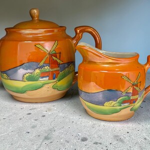 May include: A ceramic teapot and creamer set. The orange-colored set features a windmill design with red, yellow, and green accents. The teapot has a lid and spout, while the creamer has a handle and spout. The set is on a gray surface.
