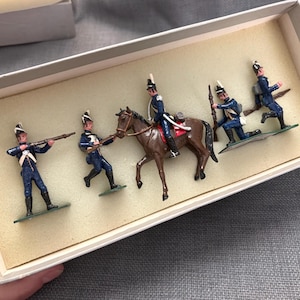 May include: A set of five painted metal toy soldiers in a rectangular box. Four soldiers wear blue uniforms with white accents and tall hats, with one soldier mounted on a brown horse. The figures are posed in various action stances with rifles.