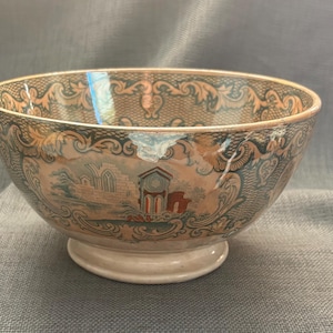 May include: A ceramic bowl featuring a detailed transfer print pattern in shades of brown, blue, and cream. The exterior displays an intricate scrollwork border surrounding a central landscape scene depicting architectural ruins and a small building.