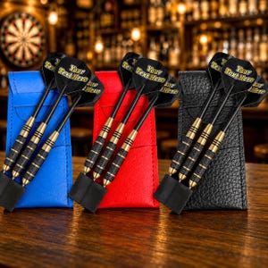 May include: Three sets of three darts with black flights featuring the text Your Text Here, black shafts, and gold-toned barrels with black textured grips. Each set is stored in a vertical leather-like case in blue, red, and black colours.