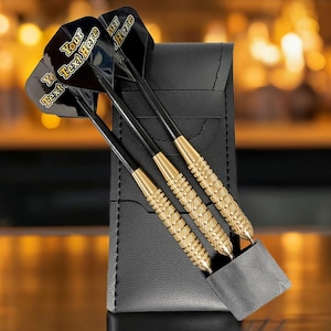 May include: Three darts with black shafts and gold-colored textured handles are displayed in a black leather case. The dart flights are black with gold text that reads "Your Text Here". The case is rectangular and has a pocket for the darts.