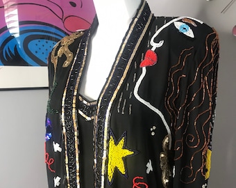May include: A black long-sleeved jacket with a colourful beaded design. The design features a stylised face, stars, and floral motifs. The jacket is made of a sheer fabric and has a V-neck.