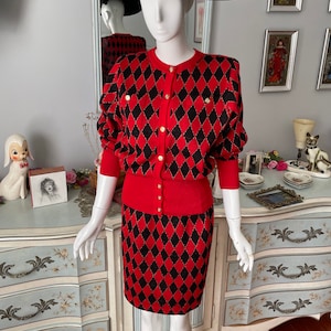 May include: A red and black knit sweater and skirt set with a diamond pattern. The set features gold buttons and a black wide-brimmed hat.