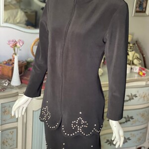 May include: A black two-piece outfit comprising a long-sleeved jacket with a high collar and a full-length zip, and a matching skirt. Both the jacket and skirt have a scalloped hemline and are decorated with small, silver-coloured studs. The outfit is displayed on a mannequin.