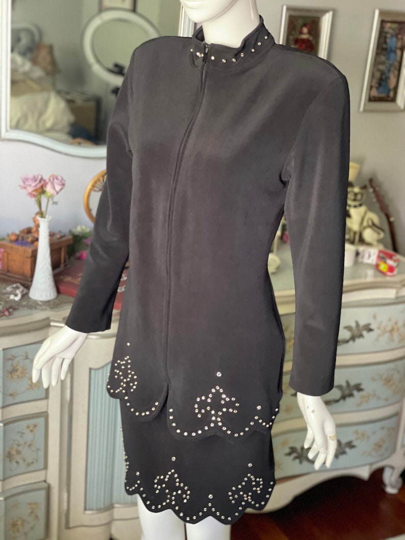May include: A black two-piece outfit comprising a long-sleeved jacket with a high collar and a full-length zip, and a matching skirt. Both the jacket and skirt have a scalloped hemline and are decorated with small, silver-coloured studs. The outfit is displayed on a mannequin.