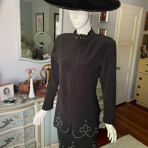 May include: A black two-piece suit and a matching wide-brimmed hat. The suit includes a long-sleeved jacket with a zip and a skirt, both decorated with sparkling embellishments. The hat is made of black velvet.