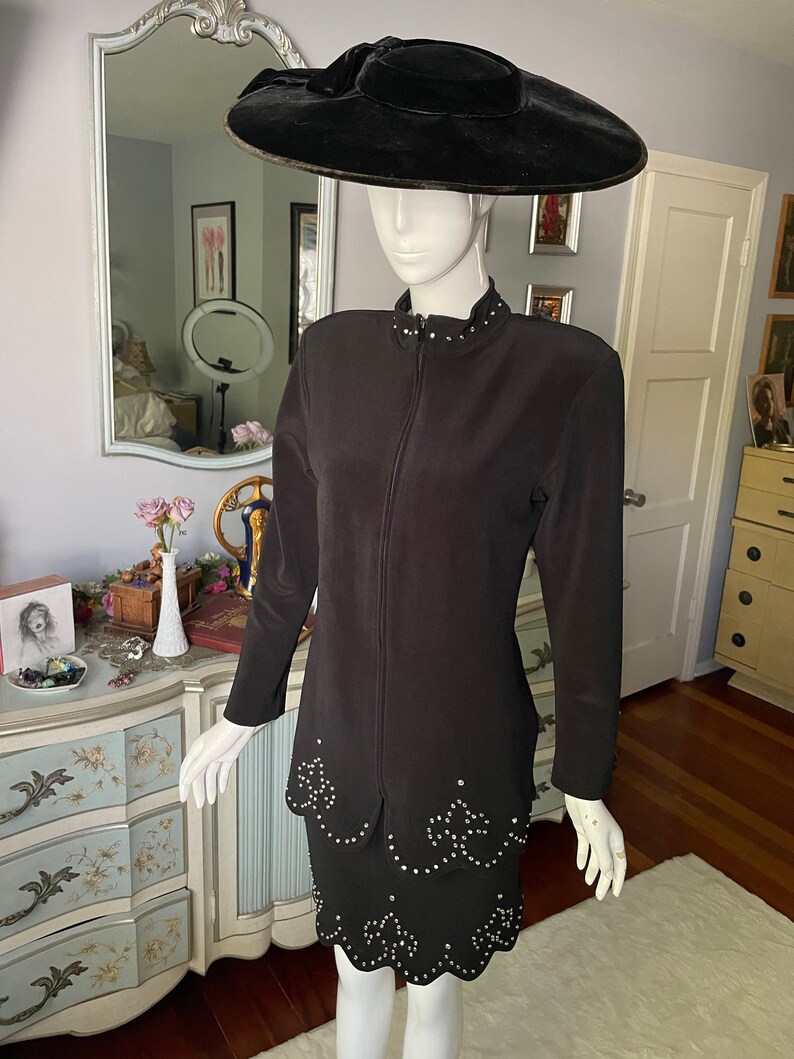 May include: A black two-piece suit and a matching wide-brimmed hat. The suit includes a long-sleeved jacket with a zip and a skirt, both decorated with sparkling embellishments. The hat is made of black velvet.