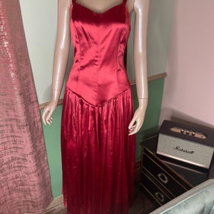 May include: A red satin dress with a sweetheart neckline and a fitted bodice. The skirt is full and flowing, with pleats throughout. The dress is sleeveless and has thin straps.