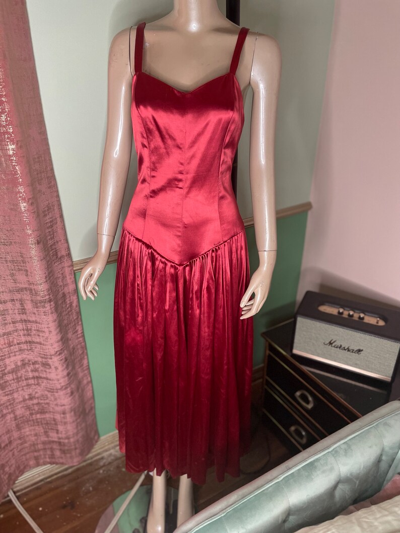 May include: A red satin dress with a sweetheart neckline and a fitted bodice. The skirt is full and flowing, with pleats throughout. The dress is sleeveless and has thin straps.