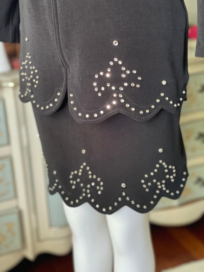 May include: Black skirt with a scalloped hem and rhinestone detailing. The skirt features a layered design with a matching top layer. Rhinestones are arranged in a floral pattern.