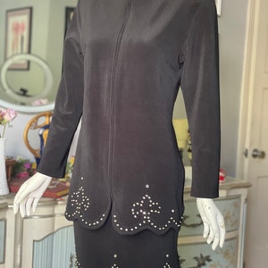 May include: A black two-piece suit comprising a jacket and skirt. The jacket has a high collar with small silver studs and a full-length zip. The skirt and jacket feature a scalloped hemline with silver stud embellishments. The suit is displayed on a mannequin.