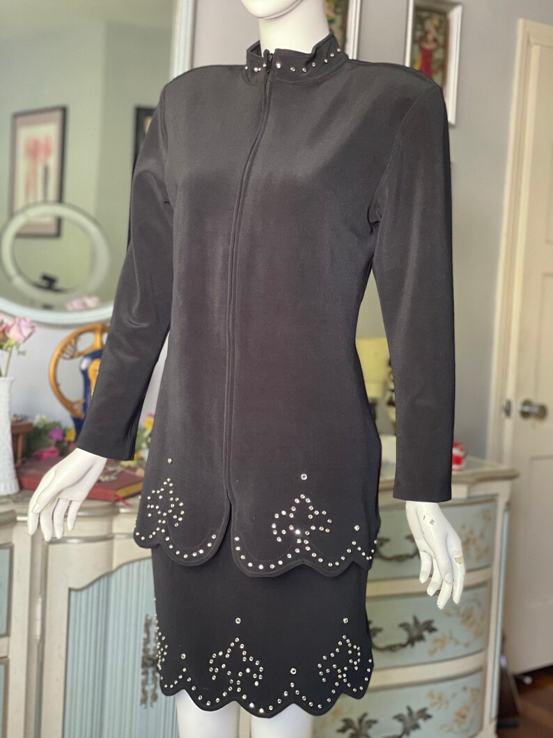 May include: A black two-piece suit comprising a jacket and skirt. The jacket has a high collar with small silver studs and a full-length zip. The skirt and jacket feature a scalloped hemline with silver stud embellishments. The suit is displayed on a mannequin.