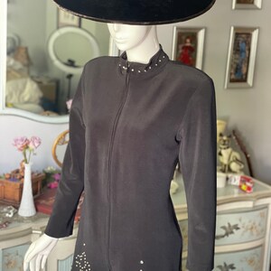May include: A black two-piece outfit on a mannequin, comprising a long-sleeved jacket with a zip and a skirt. The jacket has a high collar and is adorned with small, sparkling embellishments. A wide-brimmed black hat is on the mannequin's head.
