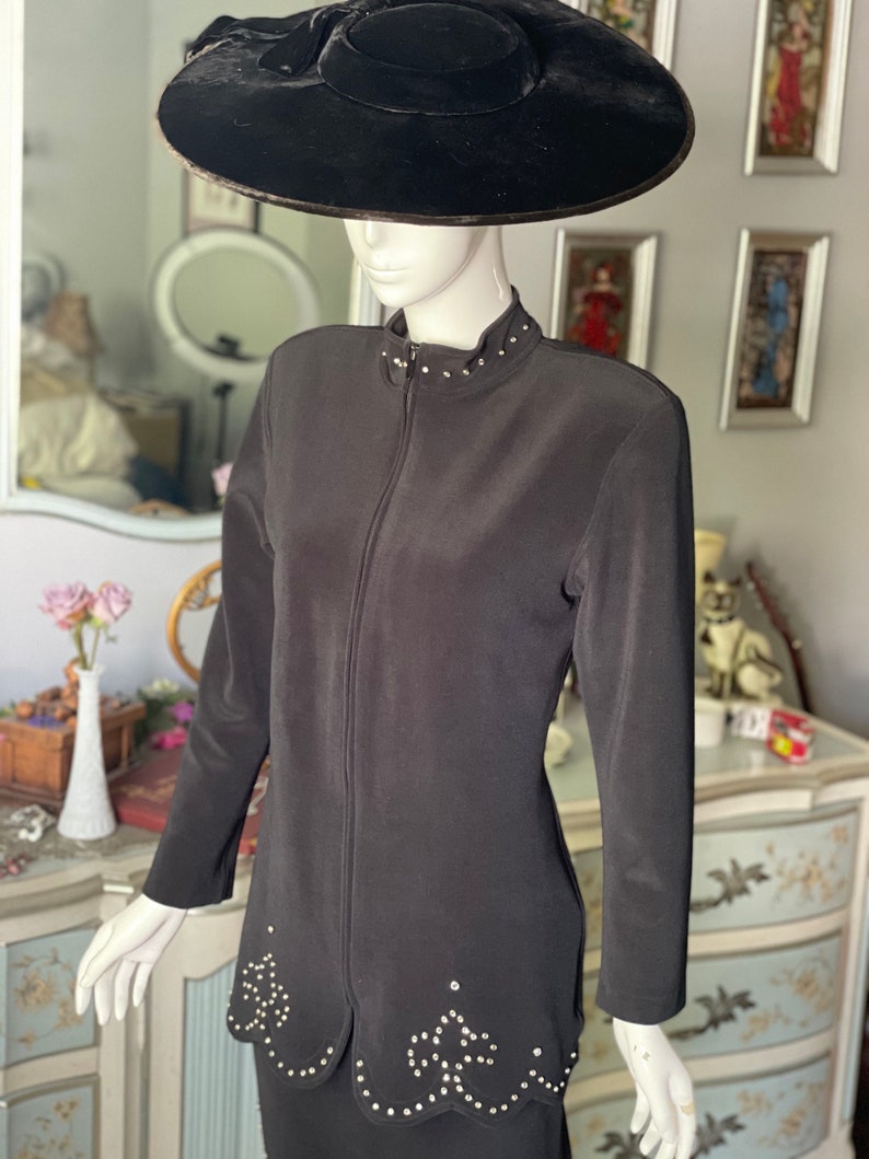 May include: A black two-piece outfit on a mannequin, comprising a long-sleeved jacket with a zip and a skirt. The jacket has a high collar and is adorned with small, sparkling embellishments. A wide-brimmed black hat is on the mannequin's head.
