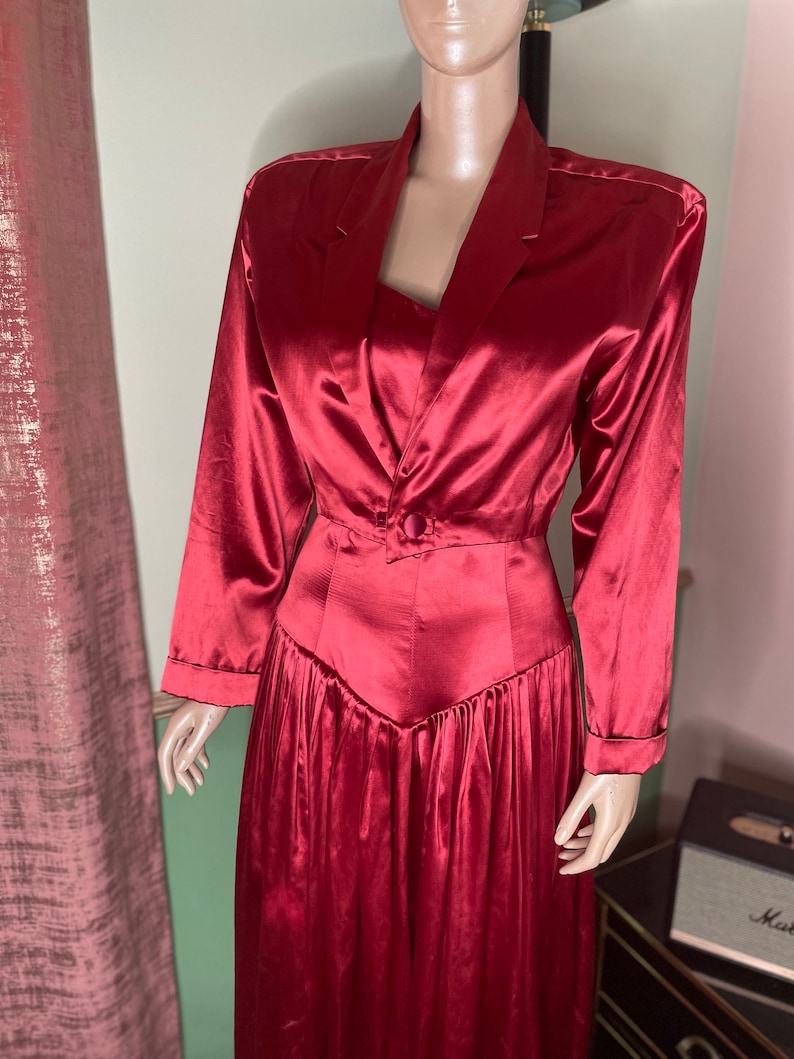 May include: A long, red satin dress with a fitted bodice and a flowing skirt. The dress has a button closure at the waist and long sleeves.