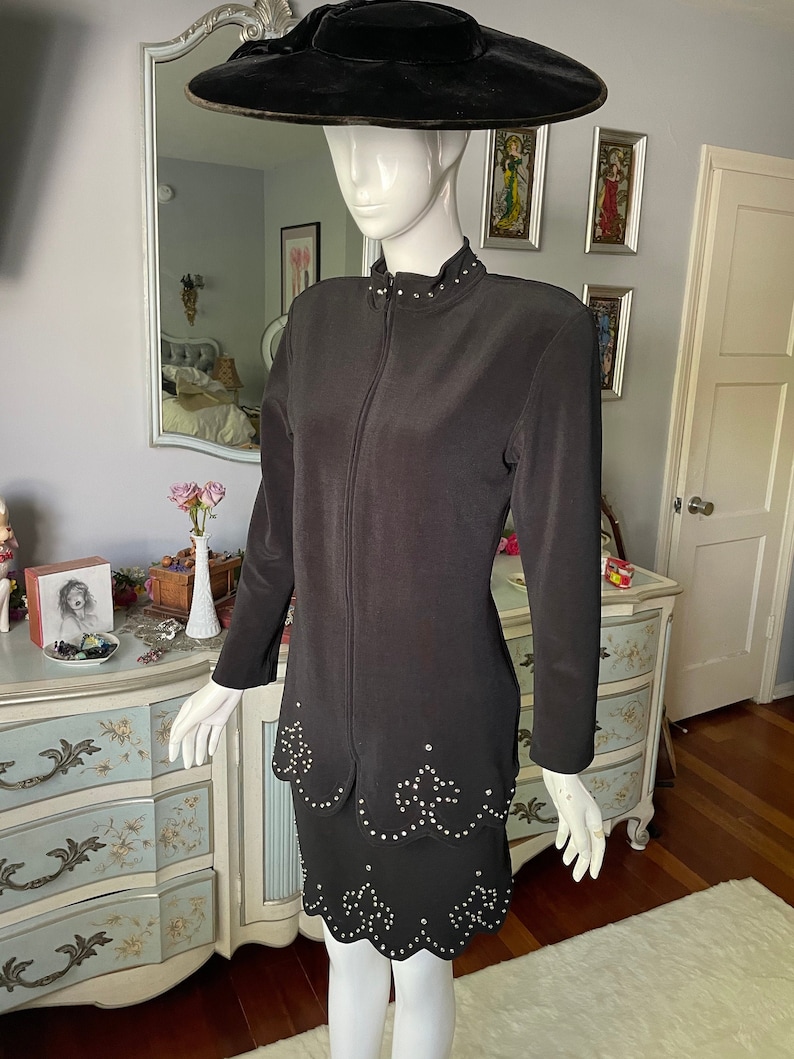 May include: A black two-piece outfit displayed on a mannequin. The jacket features a zip closure and small, round embellishments on the collar. The skirt has a scalloped hem, also embellished. A wide-brimmed black hat completes the ensemble.