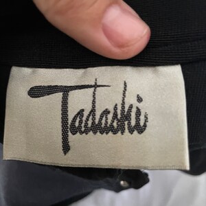 May include: A close-up shot of a black garment featuring a label with the cursive word "Tadashi". The label has a light beige background and black text. The garment is made of a dark fabric, with a few silver studs visible.