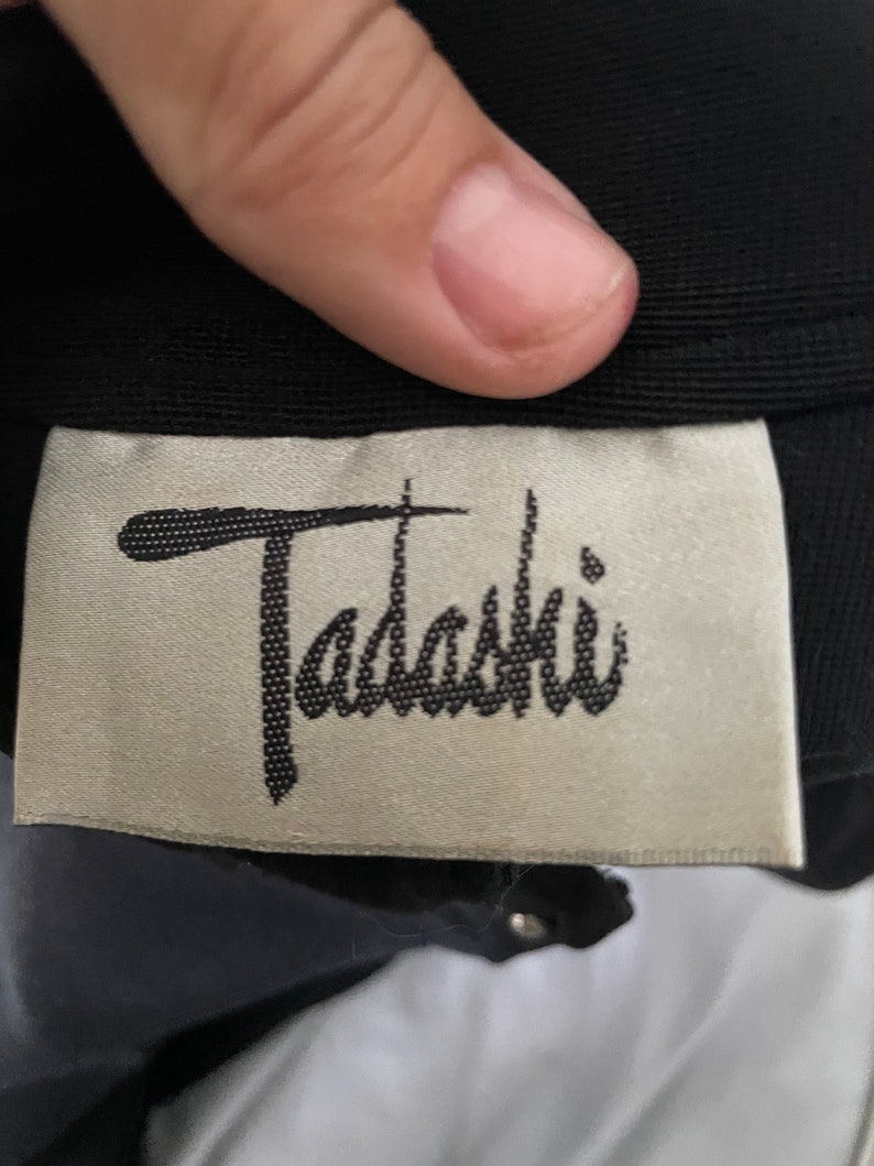 May include: A close-up shot of a black garment featuring a label with the cursive word "Tadashi". The label has a light beige background and black text. The garment is made of a dark fabric, with a few silver studs visible.