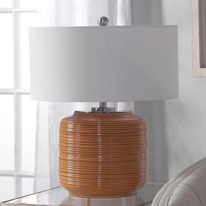 May include: A decorative table lamp with a white lampshade and a textured, orange-brown ceramic base. The base has horizontal ridges. The lamp sits on a glass-topped table. The lamp is a home decor item.